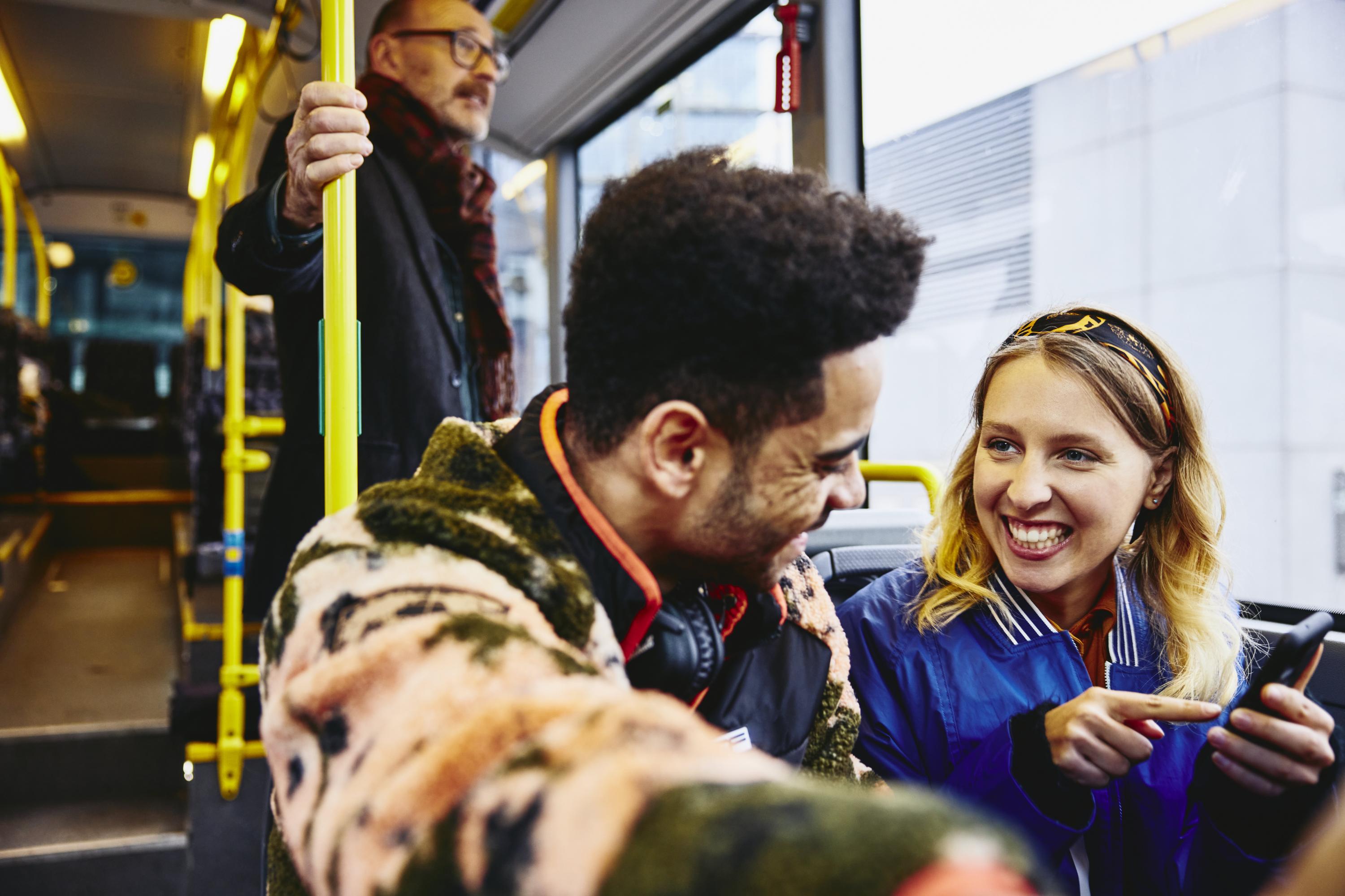 Woman and boy having a conversation, smiling, sitting in a bus. Looking at a phone. Other people in the background.