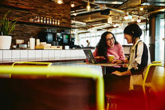 Two female sitting in restaurant having a converstation, looking at laptop, smiling.