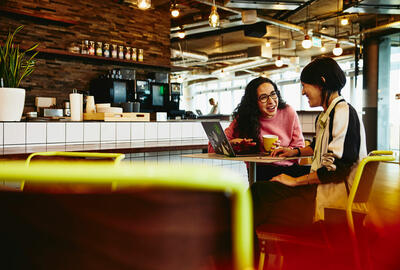 Two female sitting in restaurant having a converstation, looking at laptop, smiling.