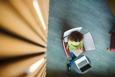 Aerial view of woman writing in a notebook with a computer next to her.